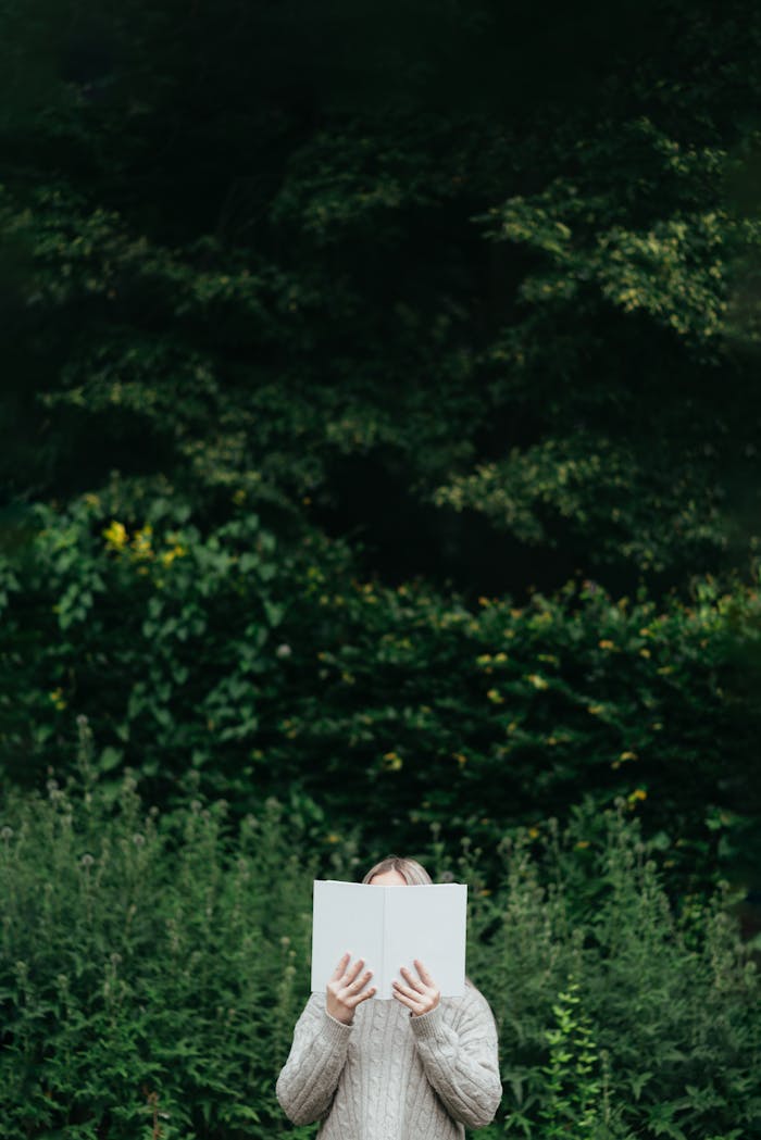 Faceless female wearing knitted sweater hiding behind book while standing in park with green foliage